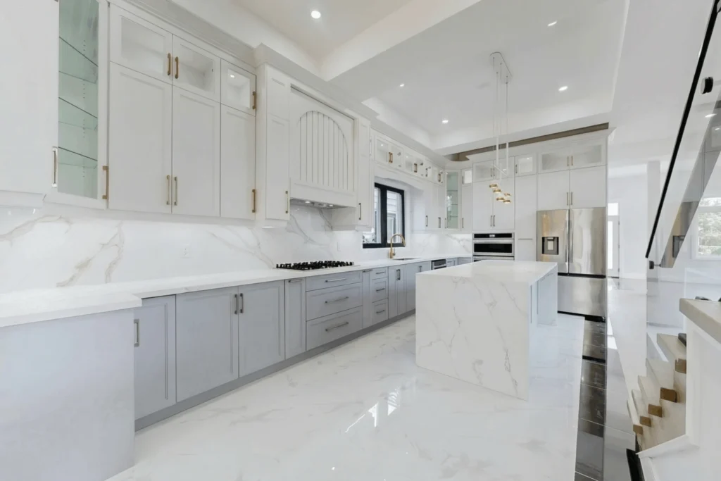 White custom kitchen with grey lower cabinets, a marble waterfall island and brass pendant lights